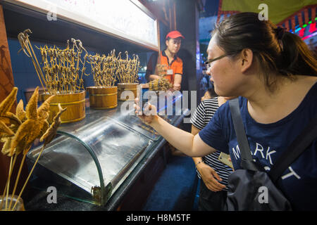 Pechino, Cina - turista femminile di scattare una foto sul suo telefono cellulare di scorpioni, cavallucci marini e stelle marine su bastoni in vendita a th Foto Stock