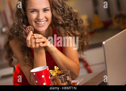 Felice giovane casalinga con spuntini di natale utilizzando laptop in cucina Foto Stock