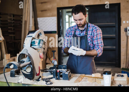 Carpenter texting qualcuno del suo smart phone vicino a sega circolare in un polveroso workshop Foto Stock