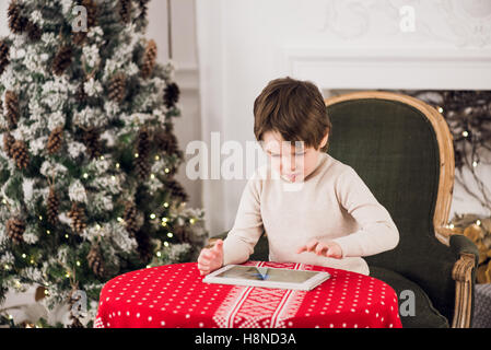 Ritratto di carino kid ragazzo seduto sulla sedia verde e giocare con i computer tablet durante il tempo di Natale. Vacanze stagioni. Foto Stock