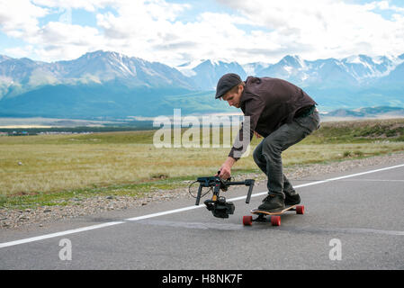 L'uomo cavalca un longboard e shoot fotocamera con electronic Steadicam. Lungo la strada nel campo sullo sfondo di montagne ghiacciai. Foto Stock