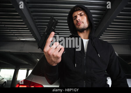 Grave giovane uomo delinquente in hoodie tenendo la pistola sul parcheggio auto Foto Stock