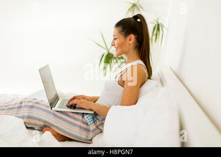 Donna sorridente recuperando i social media come lei si rilassa in letto con un computer portatile su un giorno pigro Foto Stock