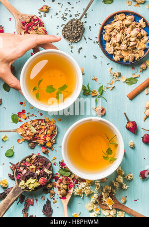 Due tazze di sano tè alle erbe con la menta, cannella, essiccato rose e fiori di camomilla in diversi cucchiai, donna di mano che regge una tazza su sfondo blu Foto Stock