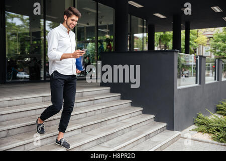 Sorridente attraente giovane imprenditore a piedi e utilizzando il telefono cellulare vicino al centro business Foto Stock