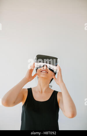 Colpo verticale della giovane donna utilizzando la realtà virtuale auricolare contro uno sfondo grigio. Felice modello femminile che indossa gli occhiali VR. Foto Stock