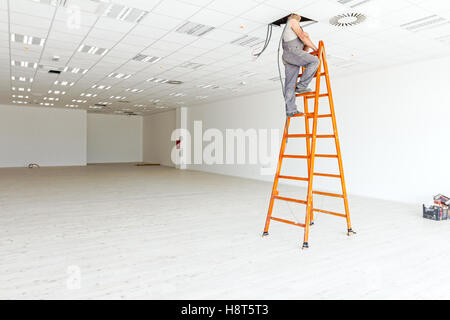Elettricista è sulla scala del peering attraverso un foro nel soffitto di un grande bianco e showroom di vuoto con pavimento in legno duro e f Foto Stock