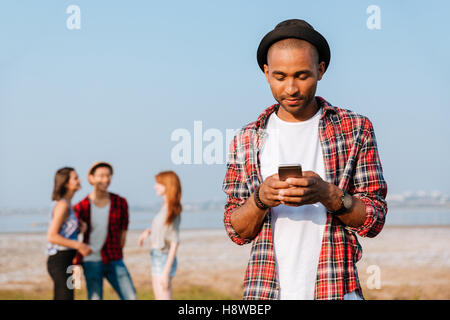 Malinconici african giovane hat in piedi vicino ai suoi amici e utilizzando il telefono cellulare Foto Stock