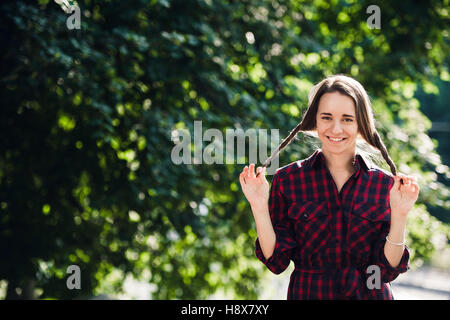 Carino ragazza adolescente in abbigliamento casual giocando con il suo trecce, guardando la fotocamera e sorridente, in piedi contro il fogliame verde Foto Stock