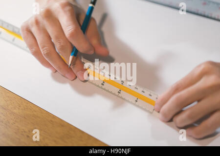 Mano di giovane donna tenendo premuto matita blu e utilizzando il triangolo misura righello sulla carta bianca messa in legno marrone tabella per la progettazione di opere. Foto Stock