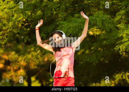 Felice ragazza adolescente in rosso dancing in natura mentre si ascolta la musica attraverso le cuffie, sorridente e guardando la telecamera su un soleggiato giorno d'estate. Foto Stock