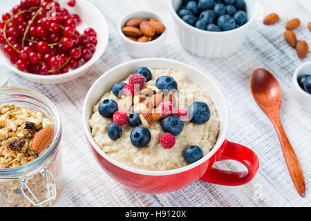 Farina di avena porridge con mirtilli freschi, lamponi, muesli e mandorle in recipiente rosso su bianco tavola. Una sana prima colazione Foto Stock