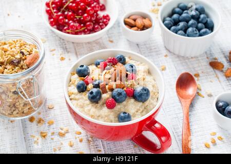 Farina di avena porridge con mirtilli freschi, lamponi, muesli e mandorle in recipiente rosso su bianco tavola. Una sana prima colazione Foto Stock