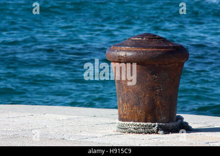 Grande vecchio arrugginito bollard su un molo di pietra, con una corda attorno ad esso e ondulato blu del mare in background Foto Stock