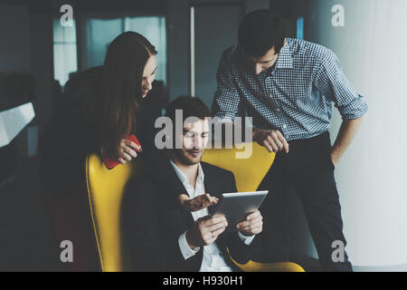 Un gruppo di giovani uomini di affari che durante la riunione: l uomo e la donna in piedi accanto alla poltrona di colore giallo con il loro capo in abito formale con tavoletta digitale, disco Foto Stock