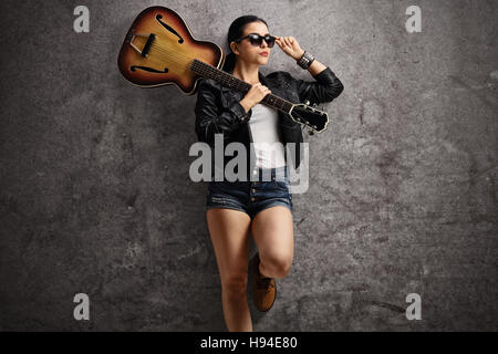 Giovane donna che indossa una giacca di pelle e occhiali da sole con una chitarra sulla sua spalla appoggiata su un arrugginito muro grigio Foto Stock