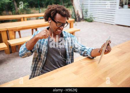 Mad irritato african giovane uomo in bicchieri e auricolari utilizzando tablet in outdoor cafe Foto Stock