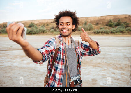 Sorridente attraente uomo africano tenendo selfie con telefono mobile e che mostra pollice in alto Foto Stock