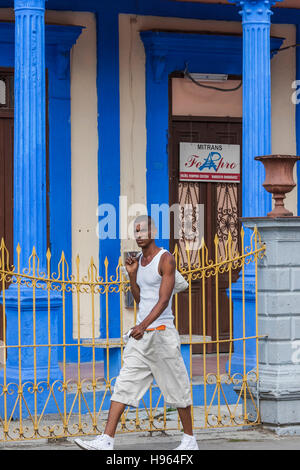 Nero giovani cubani uomo vestito di bianco cammina davanti ad un blu luminoso edificio giallo con ringhiere in Cuba Foto Stock