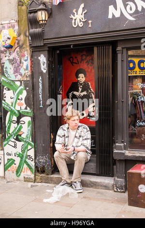 Giovane uomo seduto sulla strada di fronte a un negozio chiuso con muro coperto di graffiti e gli scarabocchi in Brick Lane, Shoreditch, Londra, Regno Unito. Foto Stock