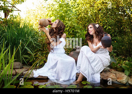 Due sorelle in tunica bianca con brocche stagno con ninfee. Una ragazza è di bere da un vecchio coccio. Foto Stock