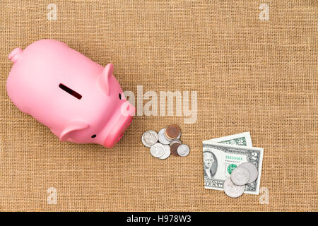 Pila di allentato in contanti di USD con dollar le banconote e le monete in euro l'alimentazione di una fame rosa salvadanaio in un concetto di risparmio, di ricchezza e di pianificazione del pensionamento in una d Foto Stock