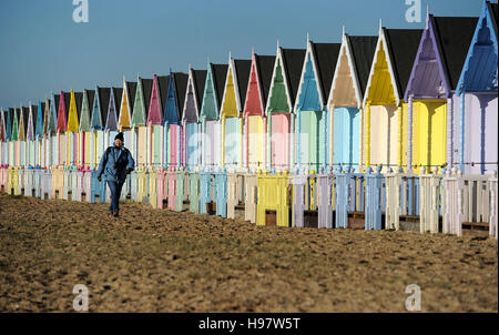 Una donna cammina davanti ad una fila di cabine sulla spiaggia, sulla spiaggia di West Mersea in Essex. Foto Stock
