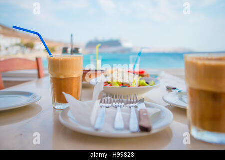 Frappé sullo sfondo della tabella e del mare grosso lainer. Estate caffè ghiacciato frappuccino in un bicchiere alto e insalata greca nel bar in spiaggia Foto Stock