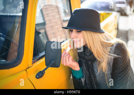 Dolce giovane donna si applica un rossetto rosso guardando lo specchio auto su una soleggiata giornata autunnale Foto Stock