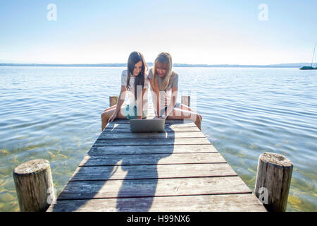 Due giovani donne di sedersi sul molo dal lago utilizzando laptop Foto Stock