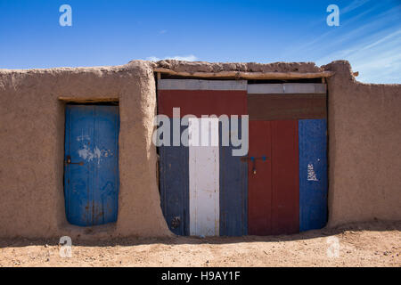 Le pareti di una casa tradizionale di creta con porta e un colorato gate. Strada di sabbia. Blu cielo con poche nuvole. Merzouga, Marocco. Foto Stock