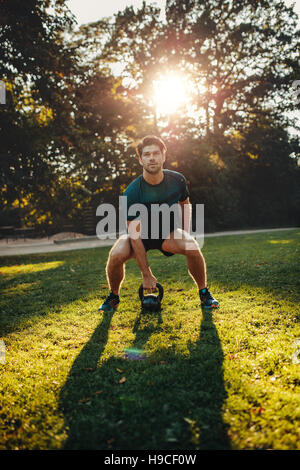 Colpo verticale di forte giovane esercitando con bollitore pesi campana nel parco. Montare e muscoloso uomo formazione presso park di mattina. Foto Stock