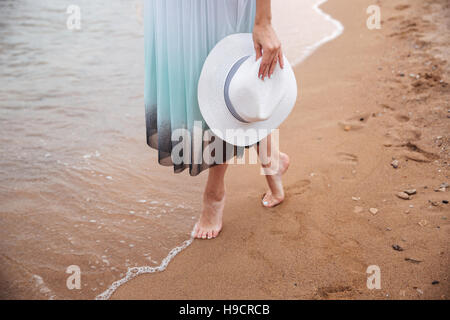 Primo piano di gambe della giovane donna in abito holding hat e correre a piedi nudi sulla spiaggia Foto Stock