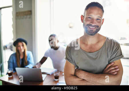 Casual cercando maschio in posa con mani incrociate su sfondo sfocato di co-lavoratori seduta a tavola in ufficio. Foto Stock