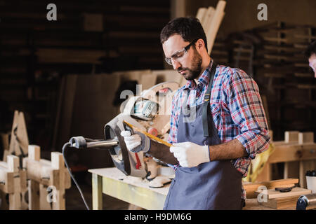 Bello il carpentiere in occhiali protettivi in piedi vicino al suo oggetto di legno in officina Foto Stock