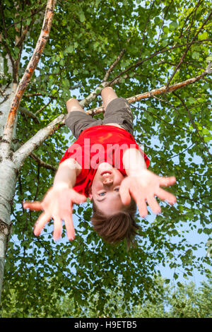 Basso angolo vista della cute teen ragazzo indossa red tshirt appeso a testa in giù da un albero guardando la fotocamera con le mani e il viso blured in movimento Foto Stock