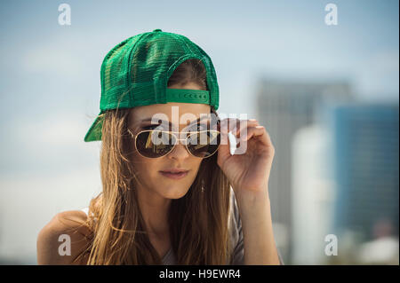 La donna caucasica indossando cappello da baseball di sbirciare da dietro gli occhiali da sole Foto Stock