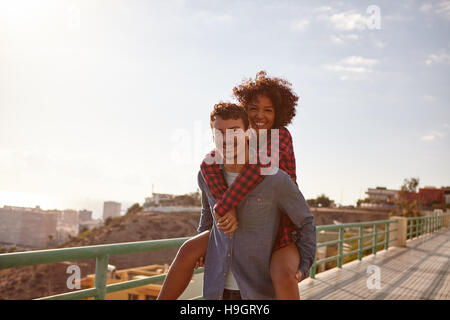 Felice giovane uomo dando la sua ragazza una piggy back come sorride un sorriso toothy con le braccia intorno al suo collo Foto Stock