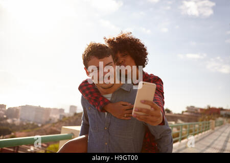 Kiss soffiaggio, piggy back riding ragazza tenendo un selfie di loro mentre è seduto sul suo fidanzato indietro tenendo con la mano destra Foto Stock