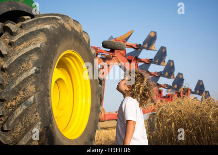 Un bambino guarda al volante di un grosso trattore con aratro sul retro in un campo di grano Foto Stock