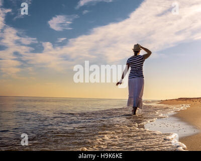 Giovane donna che indossa un cappello e mantello bianco passeggiando lungo la spiaggia verso il Rising Sun Foto Stock