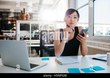 Ritratto di fiducioso donna asiatica seduto alla sua scrivania con telefono cellulare. Imprenditrice asiatici che lavorano in ufficio. Foto Stock