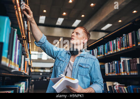 Colpo di giovani studenti di sesso maschile selezionando libro dalla libreria. Studente universitario studiare in biblioteca. Foto Stock