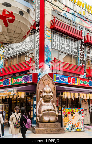 Giappone, Osaka, Shinsekai. Famosa statua Billiken, mascotte locale al di fuori su un angolo del ristorante Osaka vendita specialità alimentari, Kushi-katsu. Foto Stock