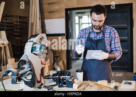 Carpenter prendendo una pausa caffè notebook di contenimento nella parte anteriore della sega circolare presso la sua officina Foto Stock