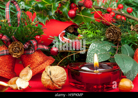 Candela rossa a forma di cuore che brucia su un tavolo decorato per natale con campane scintillanti, frutti di bosco e agrifoglio Foto Stock