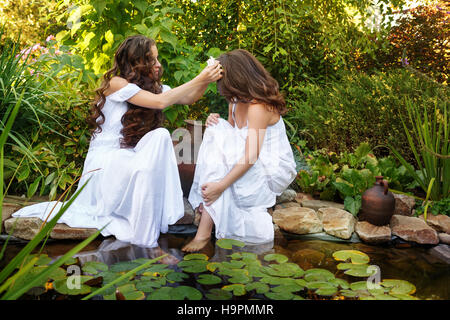 Due sorelle in bianco vestiti al laghetto. Una ragazza in fiore nei capelli tesse un altro. Foto Stock