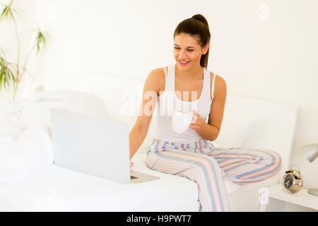 Foto di una giovane e bella donna di bere il caffè a casa nel suo letto indossando pigiami controllando il suo computer portatile Foto Stock
