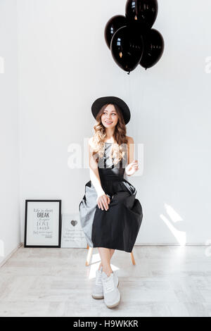 Felice carino giovane donna seduta e azienda palloncini neri su sfondo bianco con poster Foto Stock