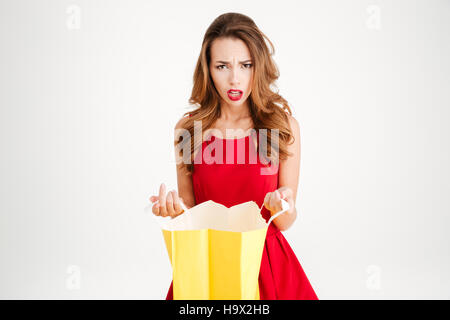 Frustrati sconvolto brunette woman in red dress holding busta gialla e guardando la telecamera isolata su uno sfondo bianco Foto Stock